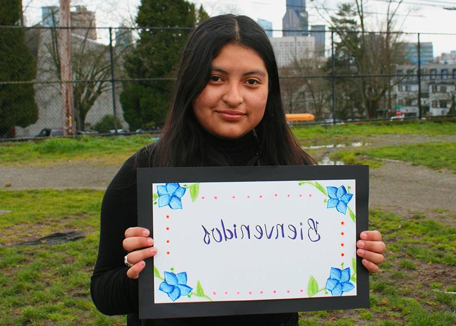 A young student poses for a photo holding a sign that says welcome in Spanish